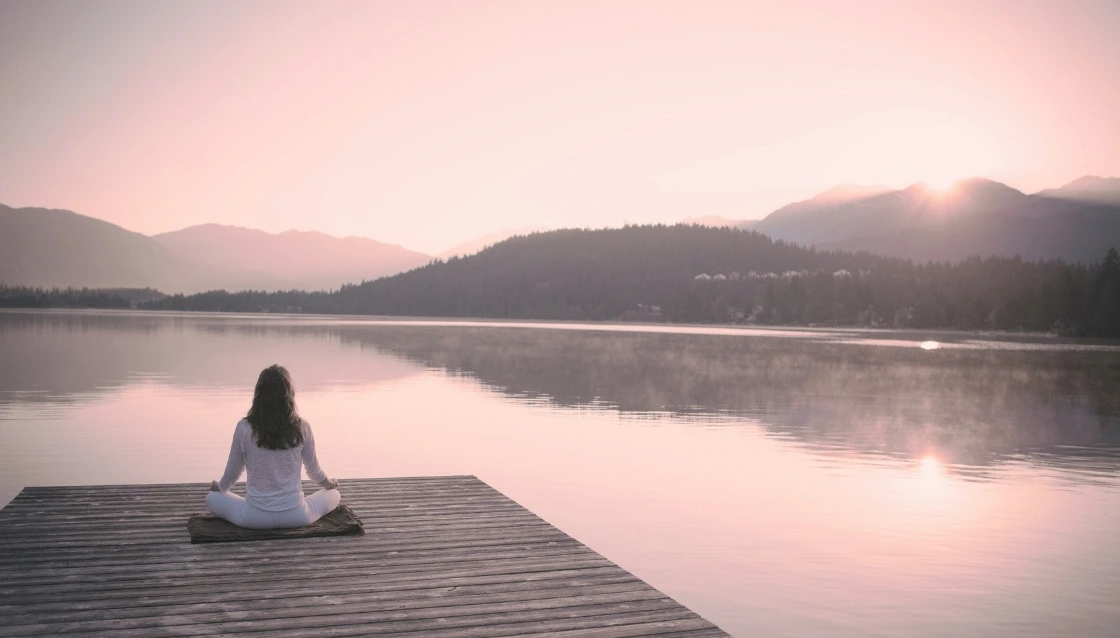 Femme assise en méditation face à un lac au lever du soleil, séance de sophrologie pour femmes dans le Var